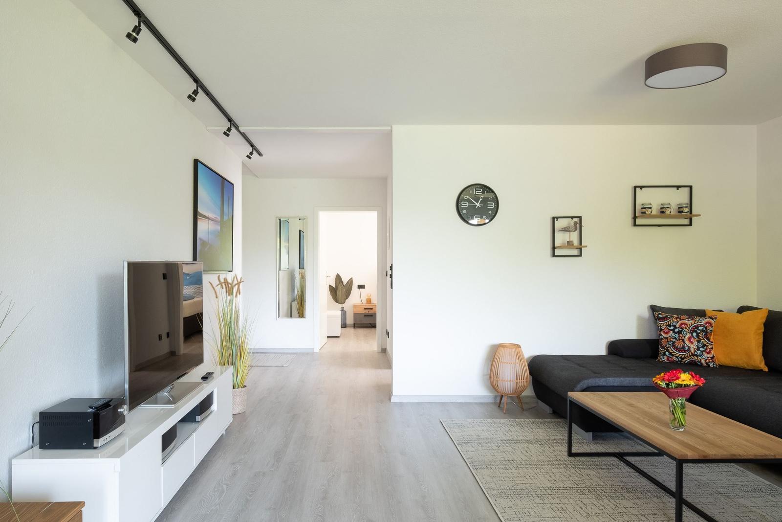 Wohnzimmer mit Sofa, Fernseher und Holztisch. Blick in den Flur mit Spiegel.