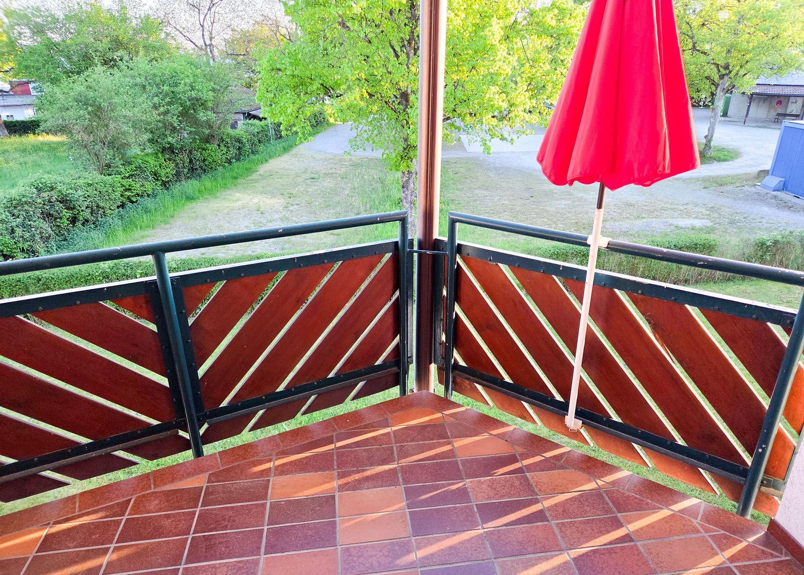 Terrasse mit rotem Sonnenschirm und Holzgeländer. Blick auf grüne Wiesen und Bäume.