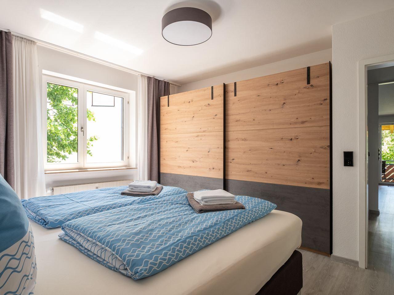 Schlafzimmer mit Doppelbett, Holzwand und Fenster mit Blick auf Bäume.