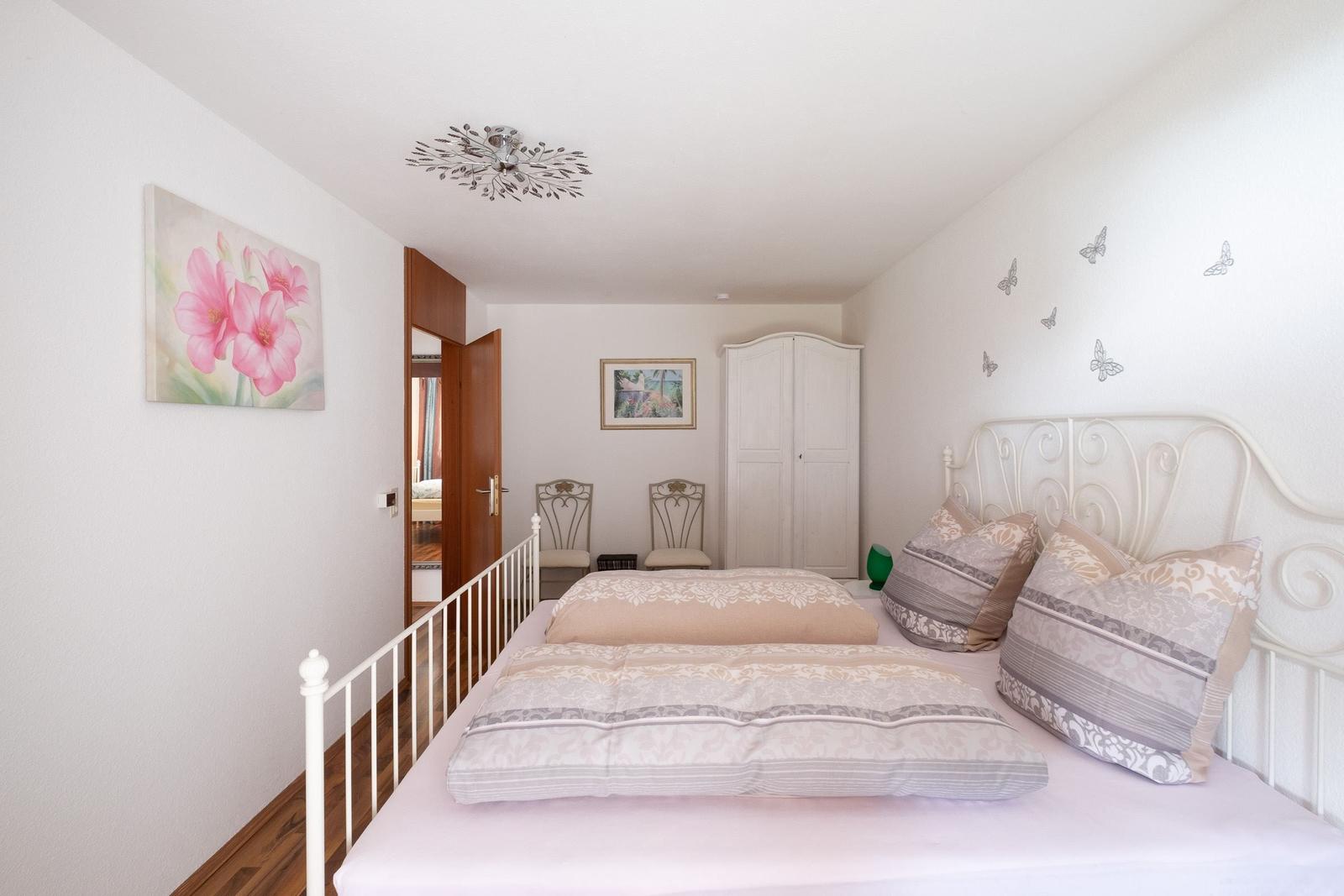 Schlafzimmer mit weißem Metallbett, Wandbild mit Blumen und Schmetterlingsaufklebern an der Wand.