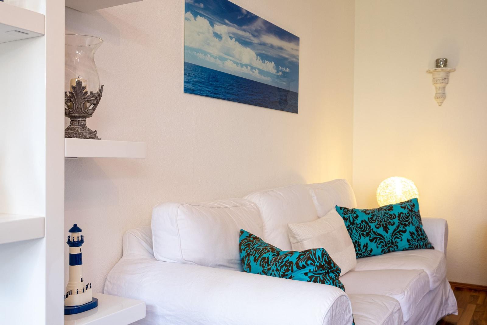 Weißes Sofa mit zwei türkisfarbenen Kissen. Wandbild zeigt Meer und Himmel. Neben dem Sofa steht eine Lampe.