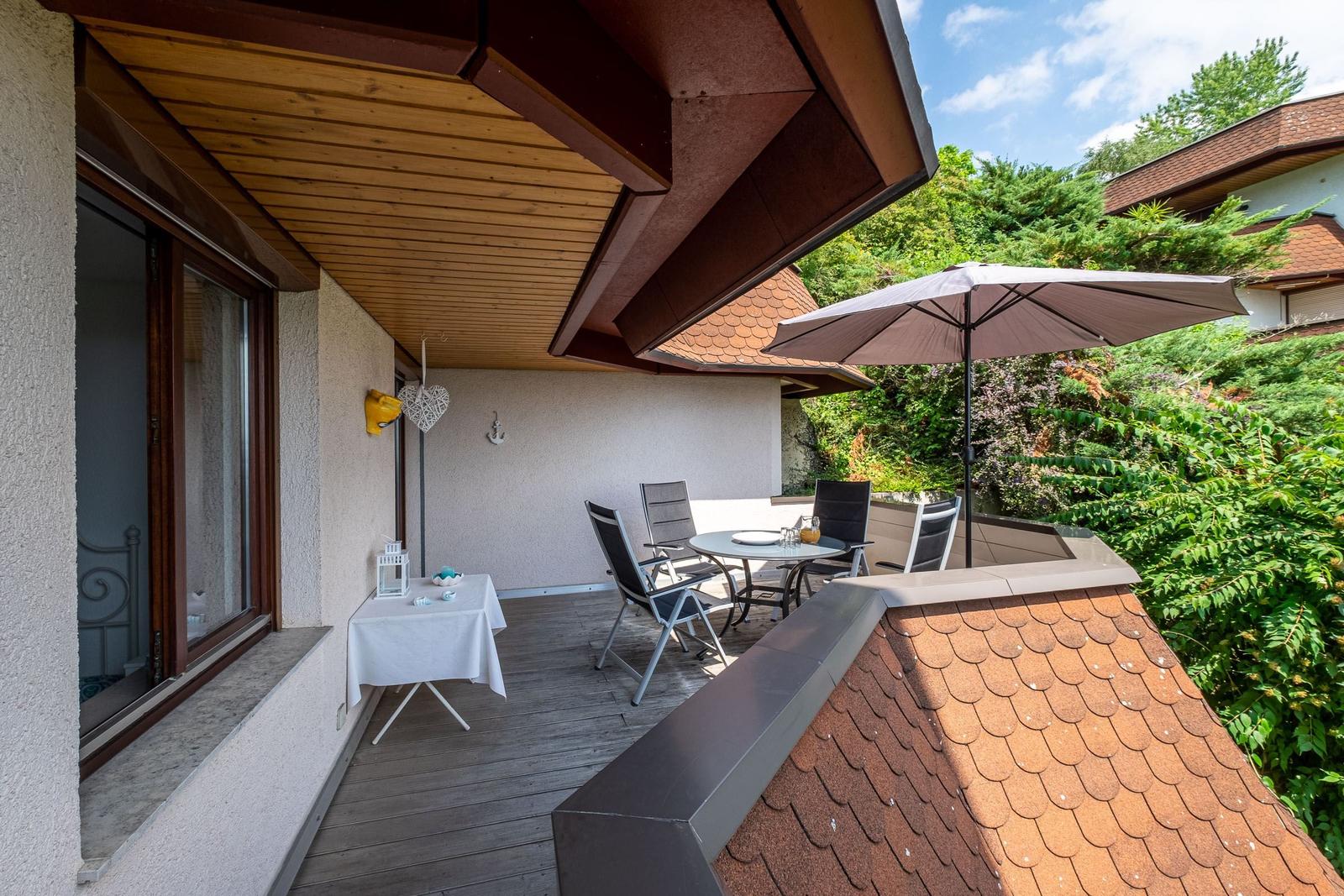 Terrasse mit Tisch, Stühlen und Sonnenschirm. Holzdecke und umliegende Grünflächen.