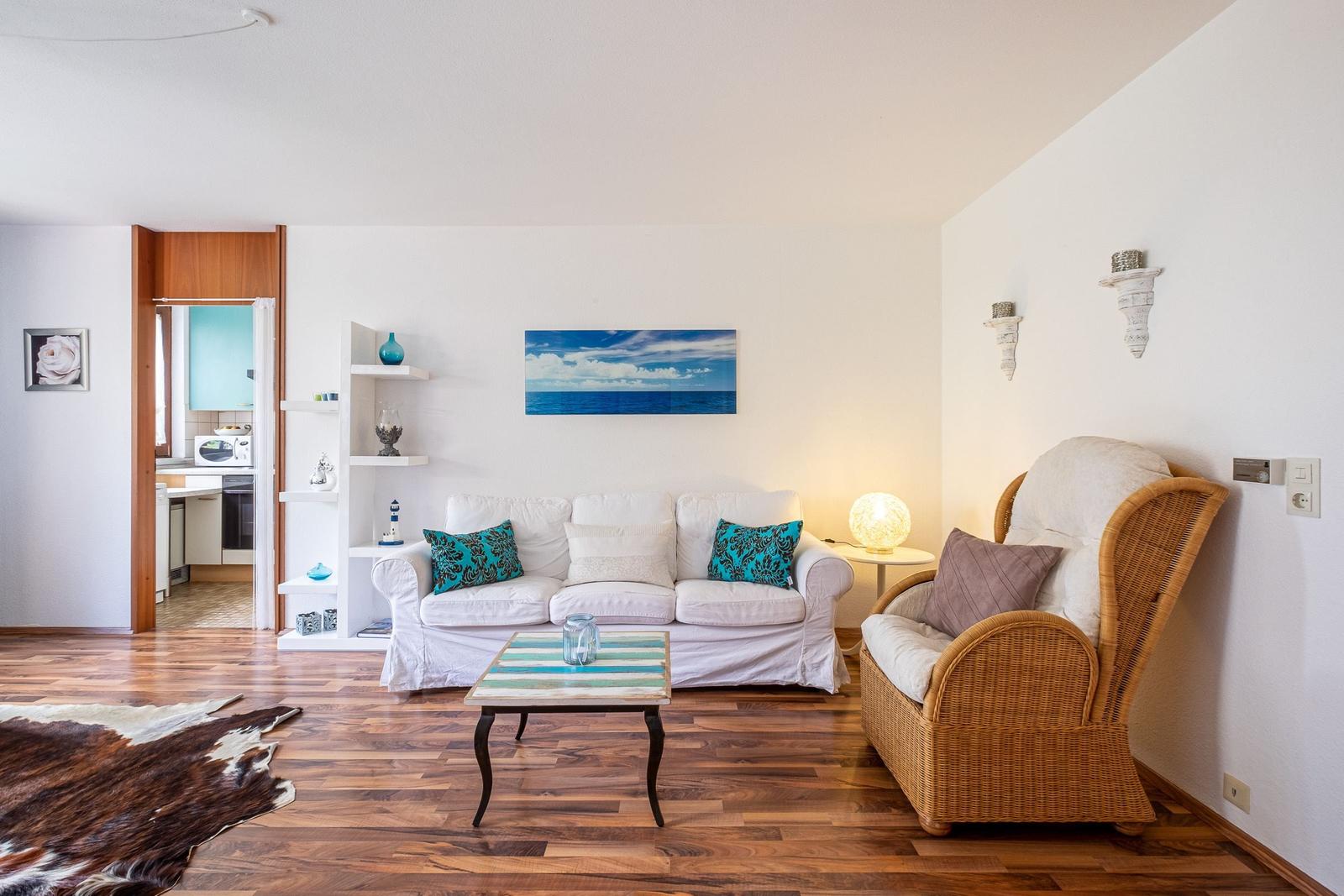Wohnzimmer mit Sofa, Sessel und Holzboden. Küche im Hintergrund sichtbar.