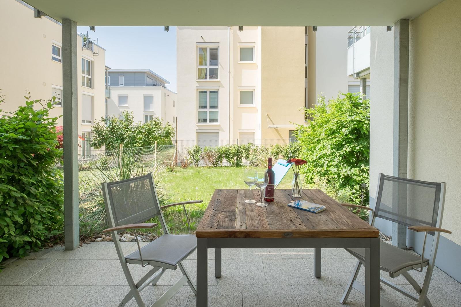 Terrasse mit Tisch und zwei Stühlen. Hintergrund: Grünfläche und Wohnhäuser.