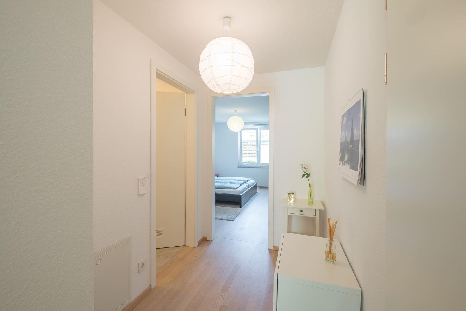 Flur mit Holzboden, weißen Wänden und Deckenleuchte. Blick in Schlafzimmer mit Bett und Fenster.