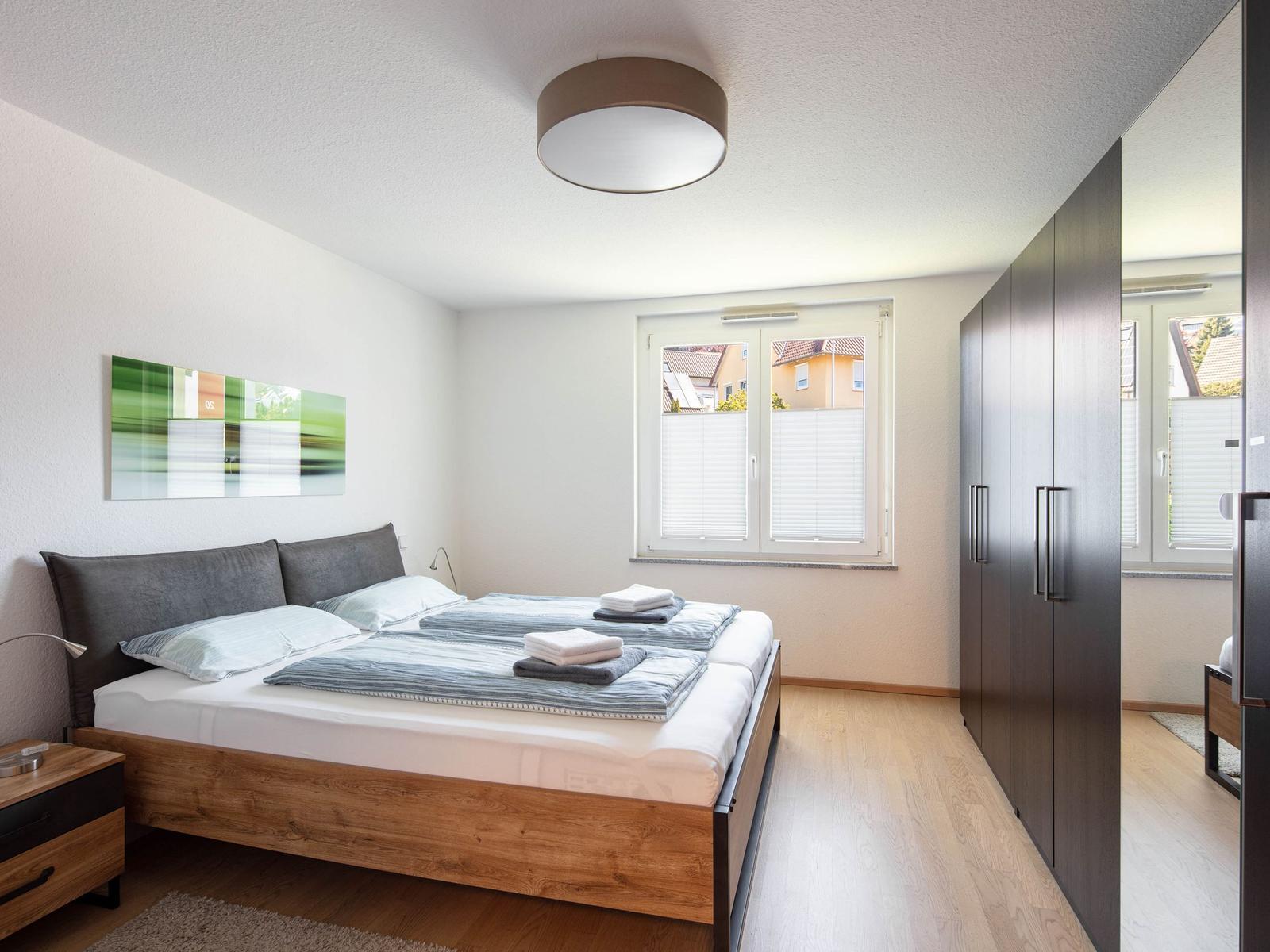 Schlafzimmer mit Doppelbett, Kleiderschrank und Fenster mit Blick auf Wohnhaus.