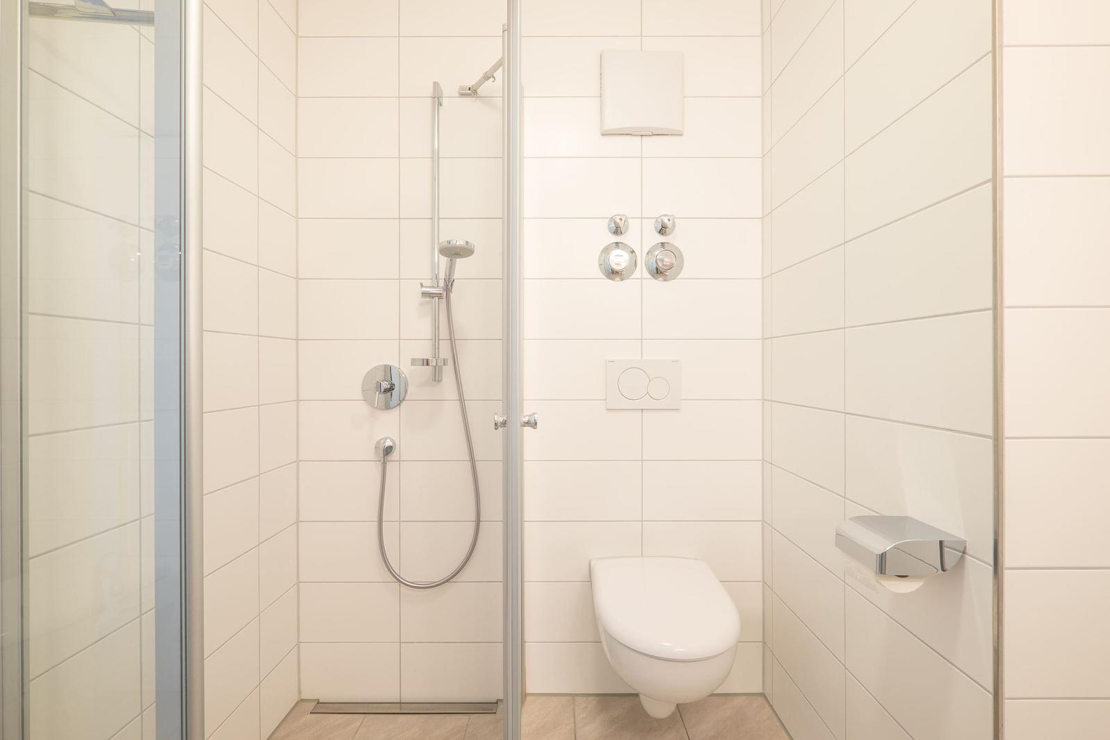 Dusche mit Duschbrause, WC und Toilettenpapierhalter in weißer Fliesenwand.