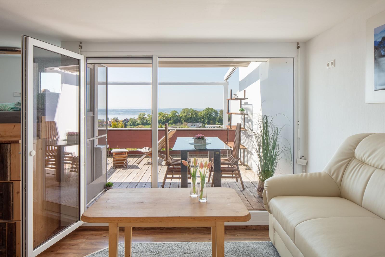Wohnraum mit Balkonblick auf Wasser und Grün. Holztisch, Sofa, Glastüren.
