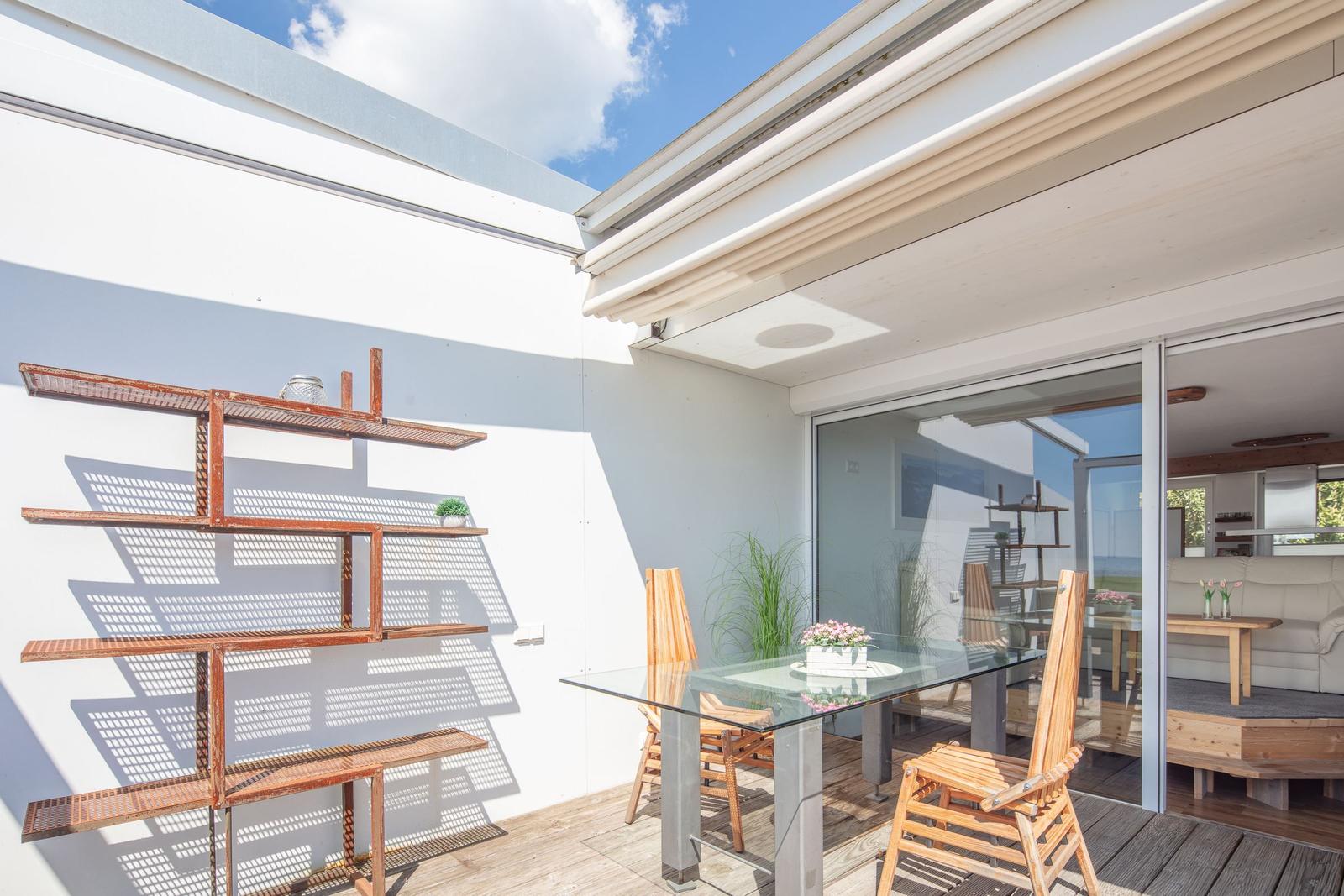 Terrasse mit Glas-Tisch, Holzstühlen und Metallregal an weißer Wand. Durchgehende Glastür zum Innenraum.