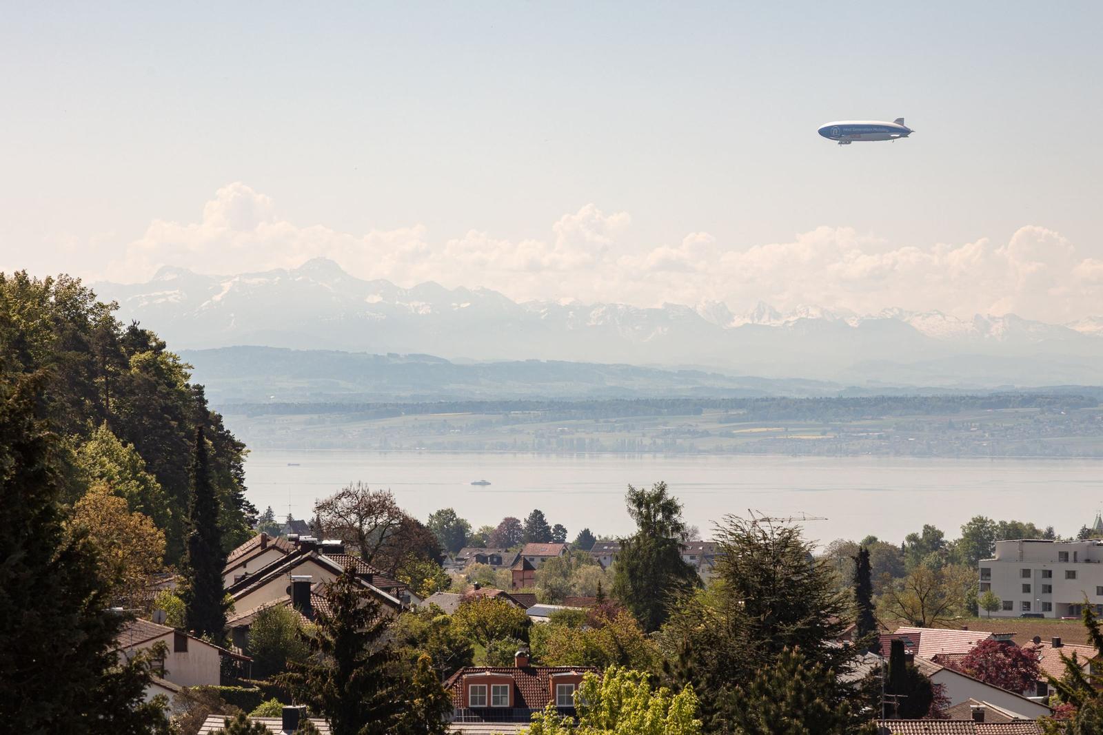 Luftschiff fliegt über See und Berge mit Blick auf Wohnhäuser.