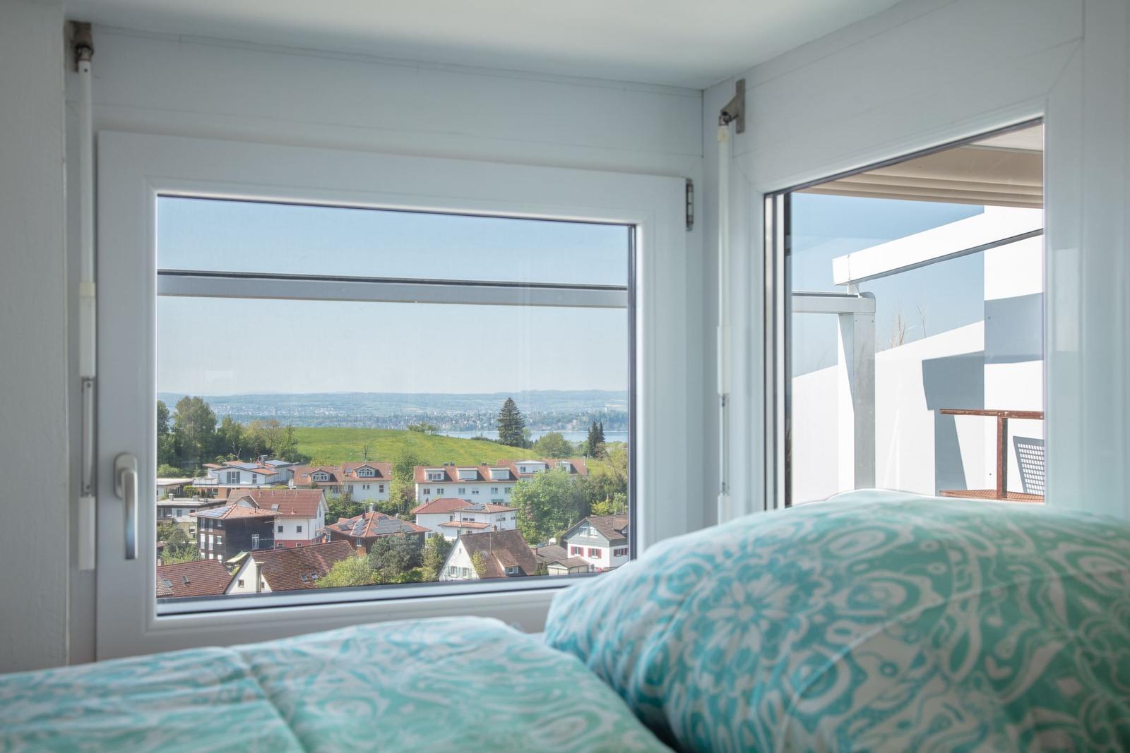 Zimmer mit Blick auf grüne Hügel und Häuser. Durchgehendes Fenster und Balkontür.