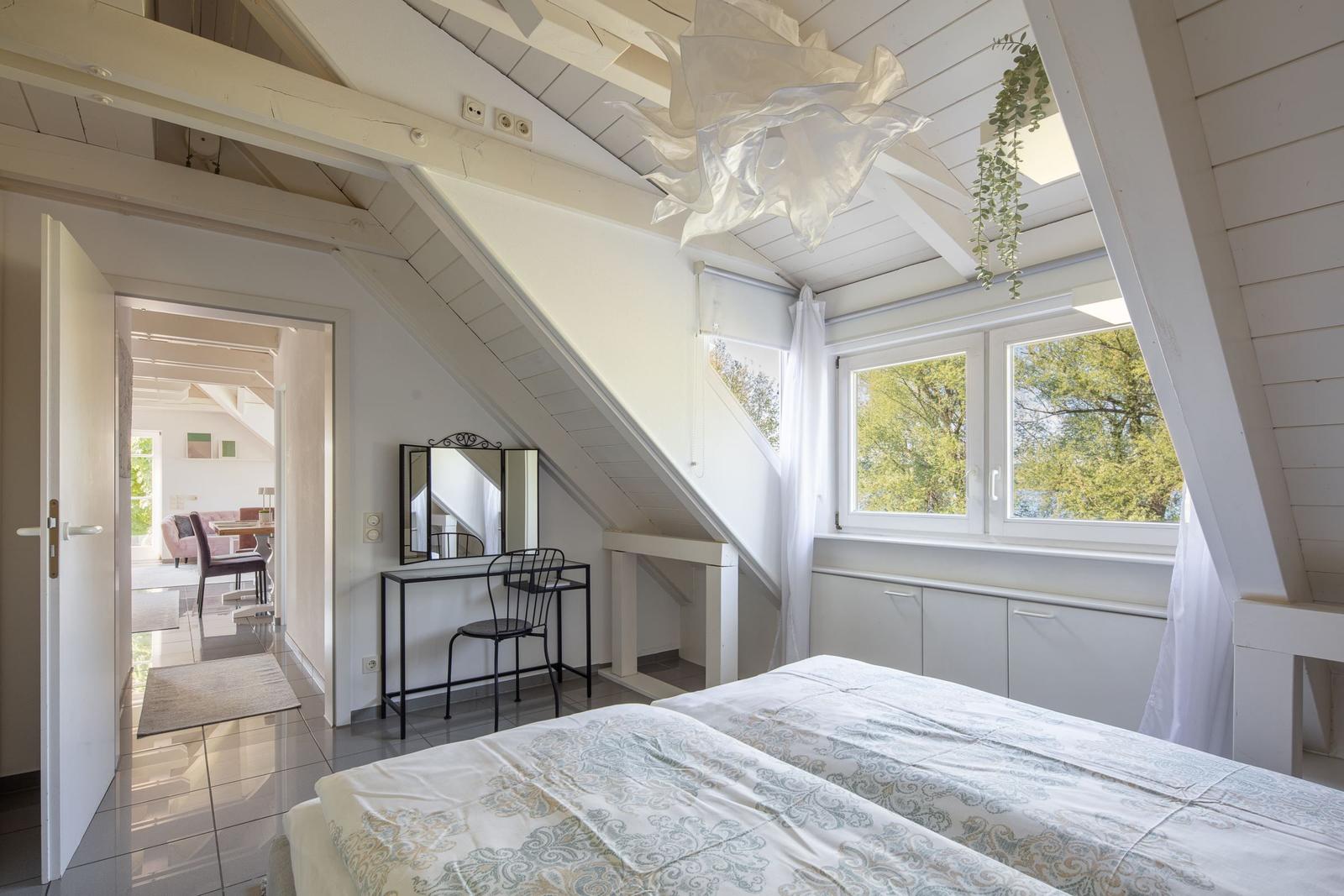 Schlafzimmer mit Dachschrägen, Fenster und Schminktisch. Blick in das angrenzende Wohnzimmer.