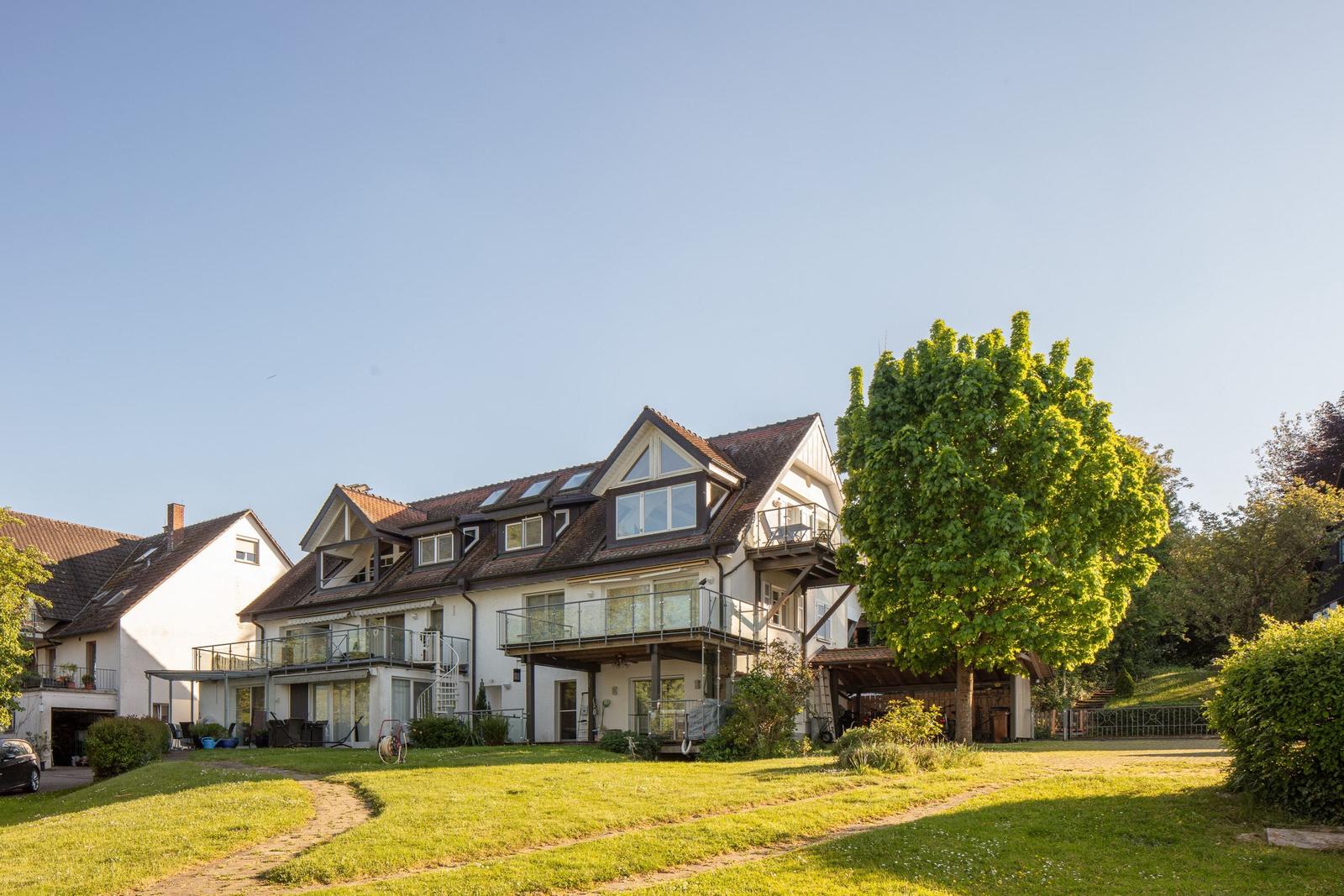 Ein zweistöckiges Haus mit Balkonen und einem großen Garten. Ein großer Baum steht im Vordergrund.
