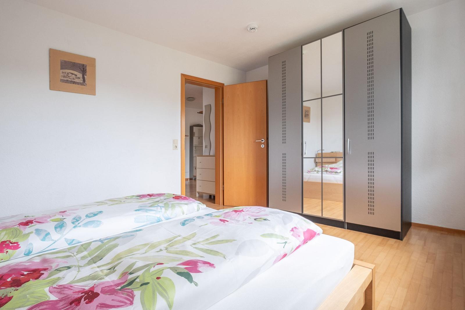 Schlafzimmer mit Bett, Spiegelschrank und Holzboden. Tür führt in angrenzenden Raum.