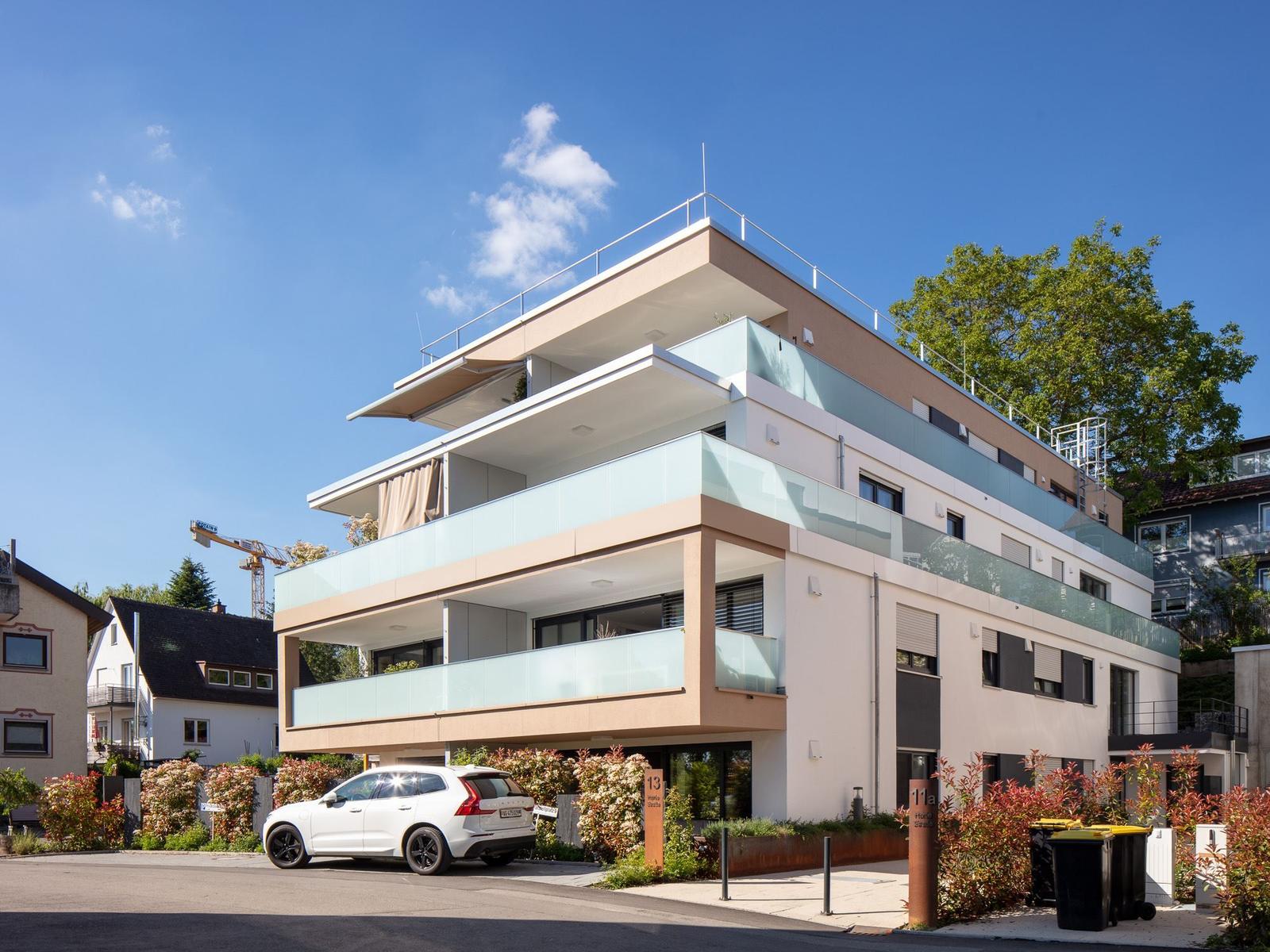 Moderne Mehrfamilienhaus mit Balkonen und Terrasse. Ein weißer Wagen parkt vor dem Gebäude.