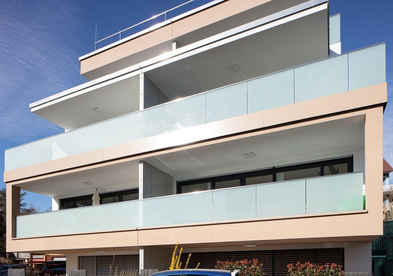 Moderne Wohnung mit drei Balkonen und Glasgeländern unter blauem Himmel.