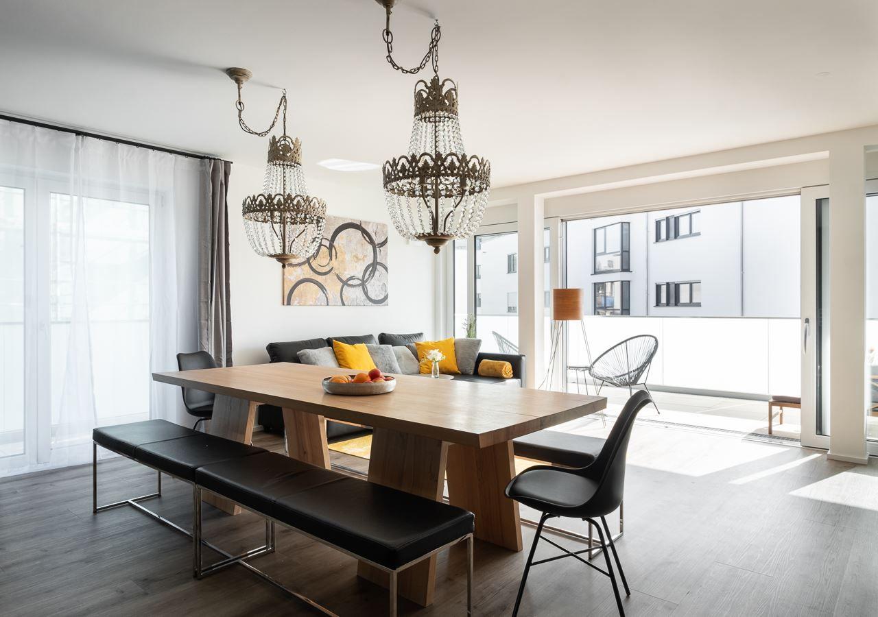 Essbereich mit Holztisch, schwarzen Bänken und zwei Kronleuchtern. Wohnbereich mit Sofa und Balkon.