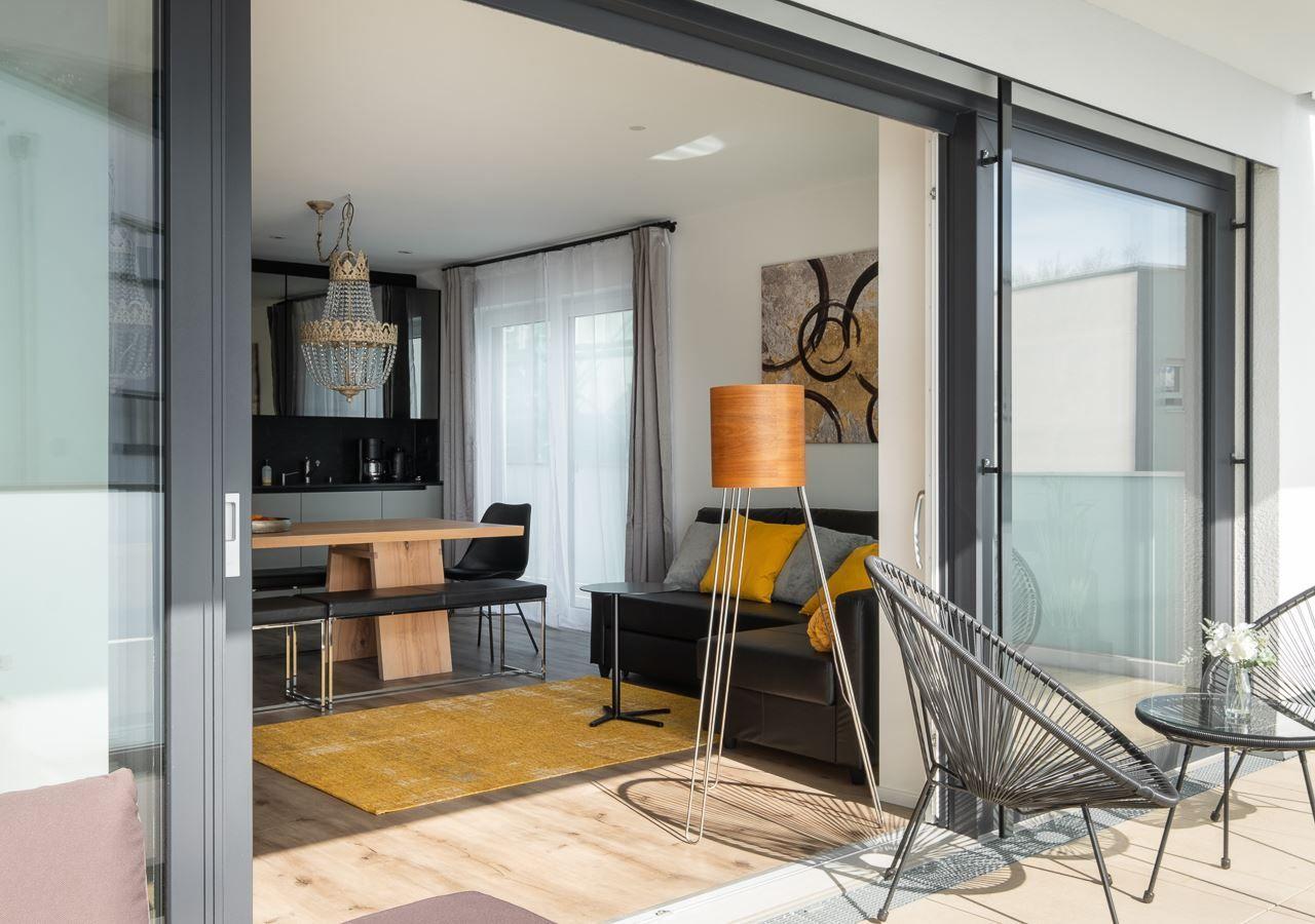 Wohnbereich mit Sofa, Stehlampe und Terrasse mit Stühlen. Durchgehende Glasfront.