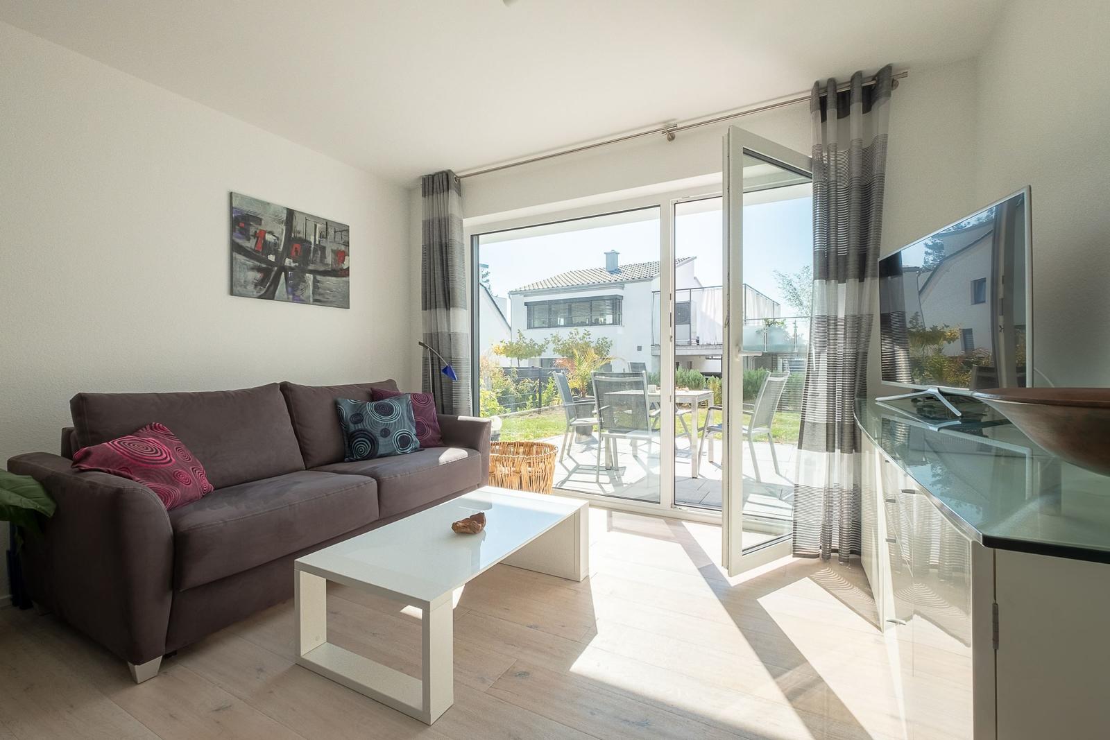 Wohnzimmer mit Sofa, Couchtisch und TV. Durchgang zur Terrasse mit Gartenmöbeln.