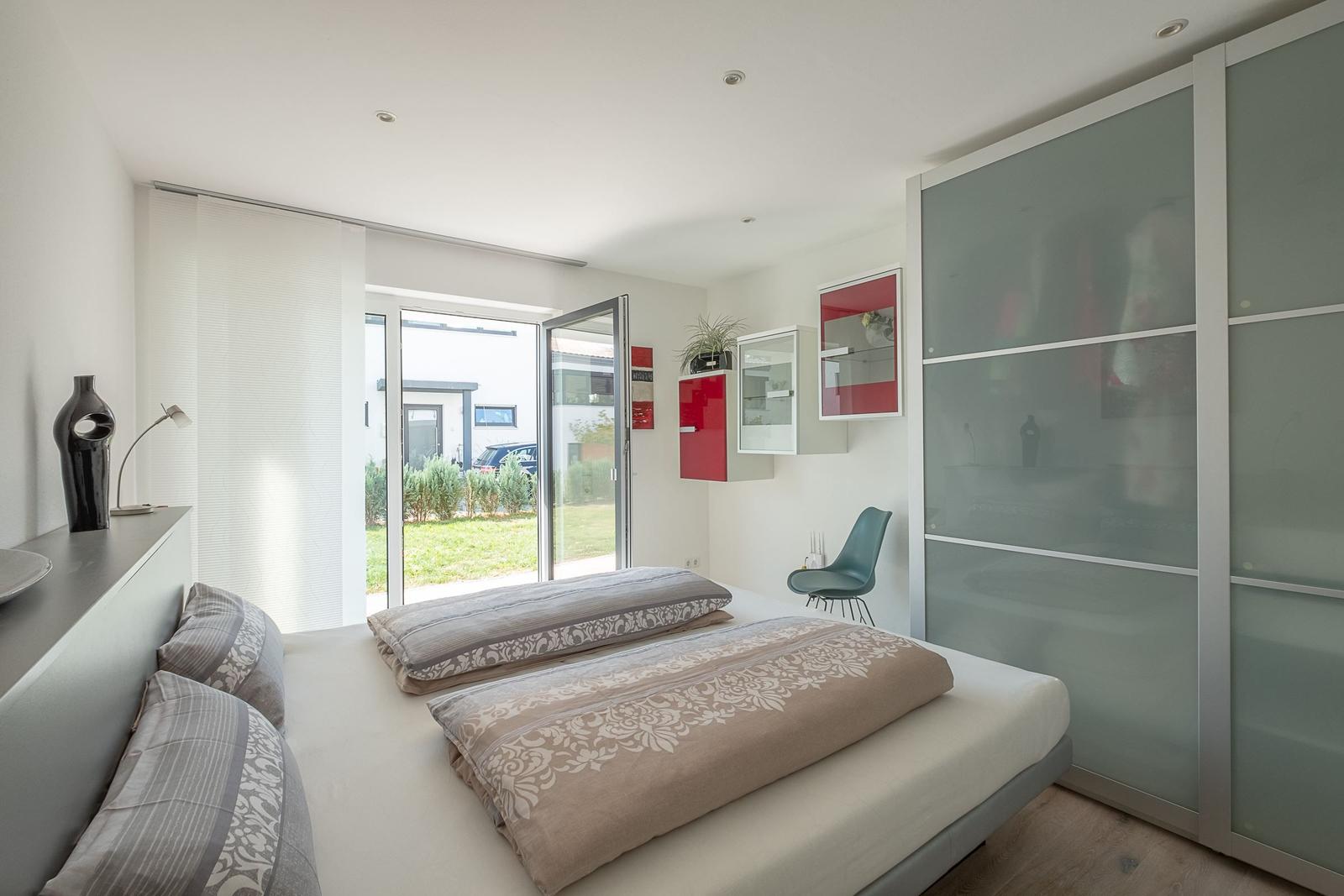Schlafzimmer mit Bett, Schrank und Glasfront. Durchgang zur Terrasse mit Blick auf Garten.