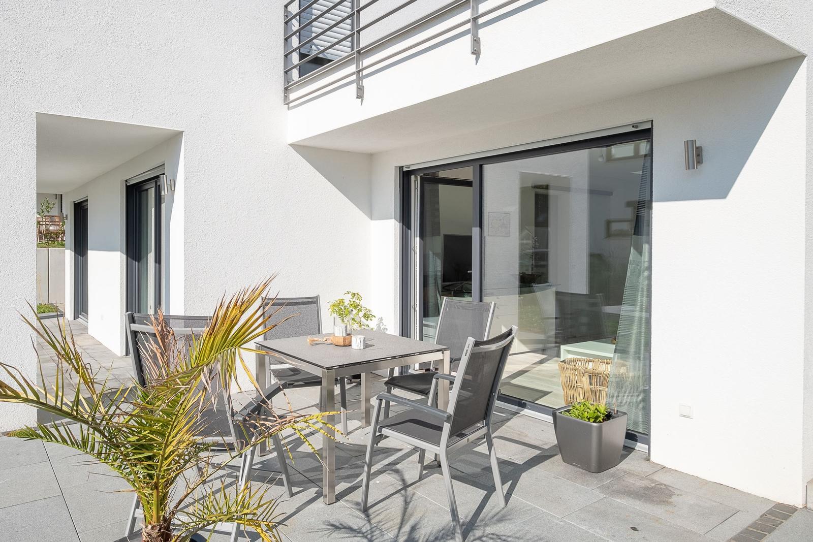 Terrasse mit Tisch, Stühlen und Palme. Glasfront öffnet zum Innenraum.