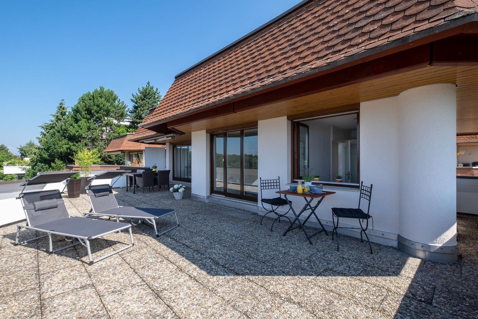 Terrasse mit Liegestühlen und Tisch. Haus mit rotem Dach und weißen Wänden.