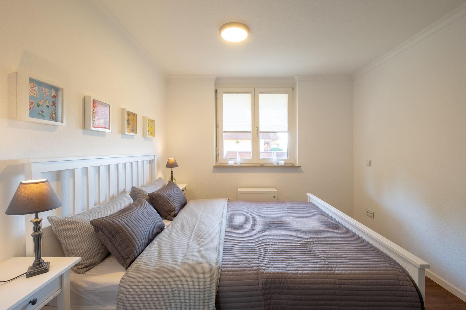 Schlafzimmer mit weißem Bett, Fenster und Nachttisch mit Lampe.