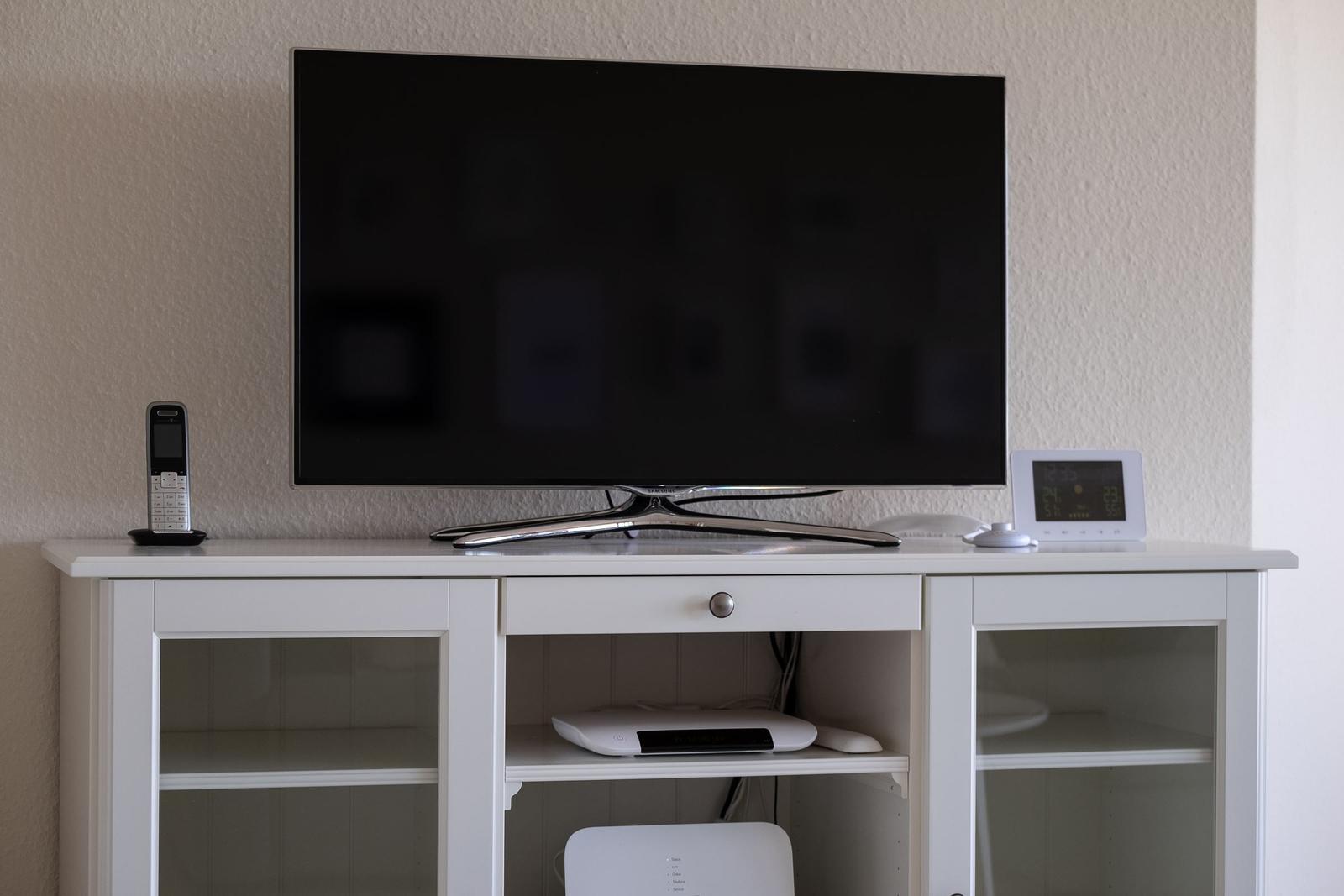 Flachbildfernseher auf weißem Sideboard mit Telefon und Digitaluhr.