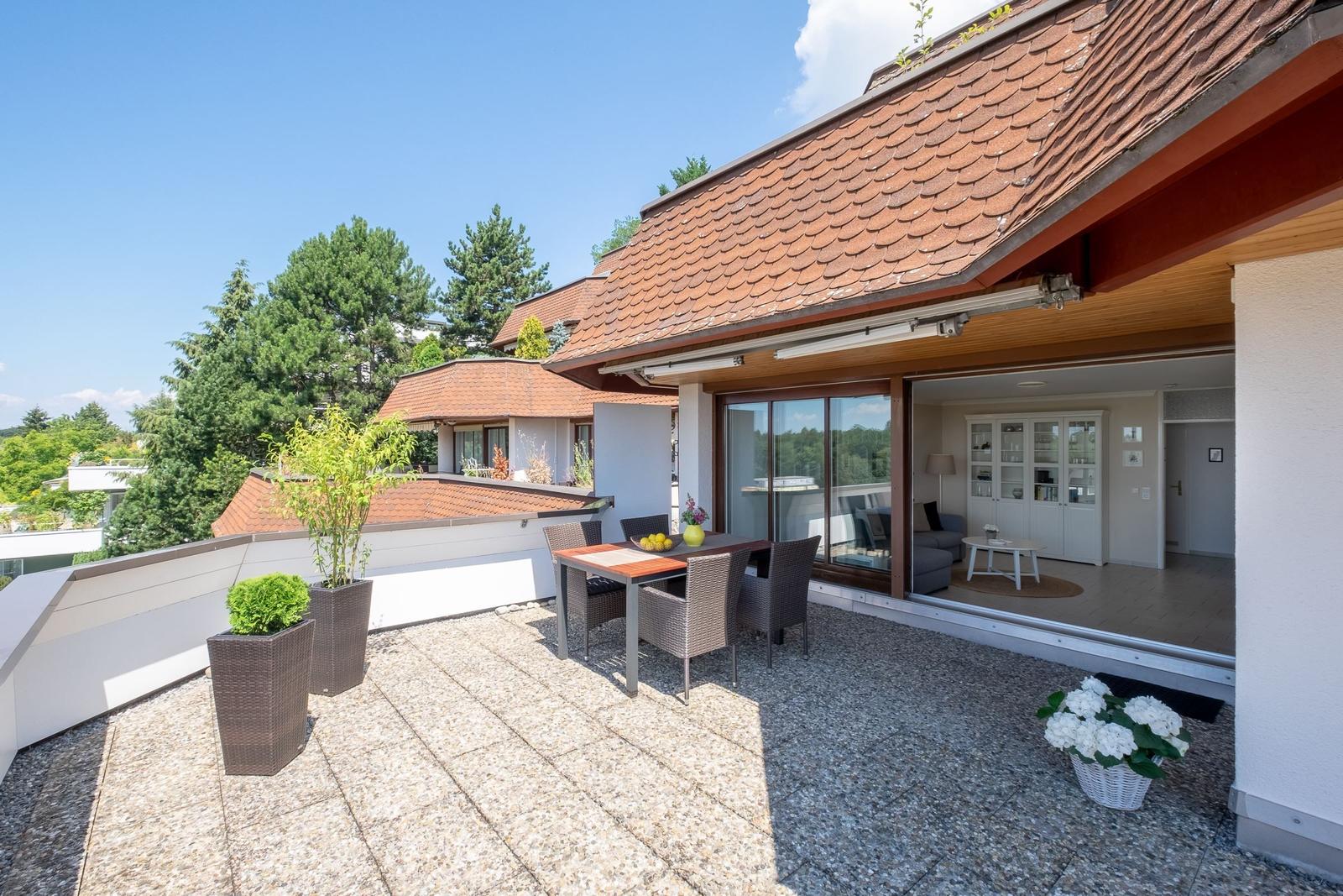 Terrasse mit Tisch, Stühlen und Pflanzen. Blick ins Haus mit Wohnzimmer und Glasfront.