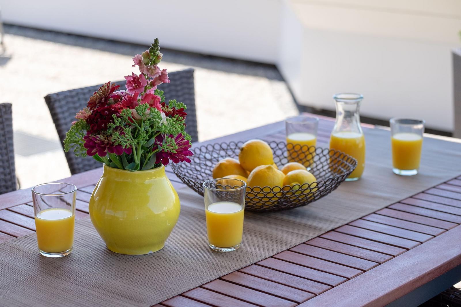 Außentisch mit Blumen, Orangensaft und Zitronen. Holzplatte und Stühle sichtbar.