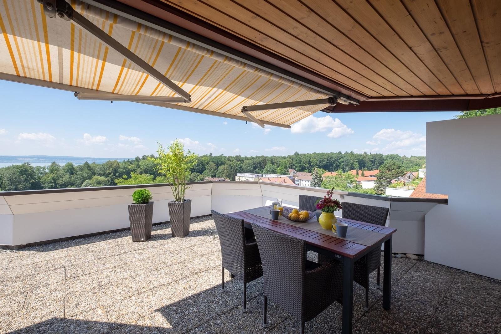 Terrasse mit Sitzgruppe, Sonnenschutz und Blick auf Wald und See.