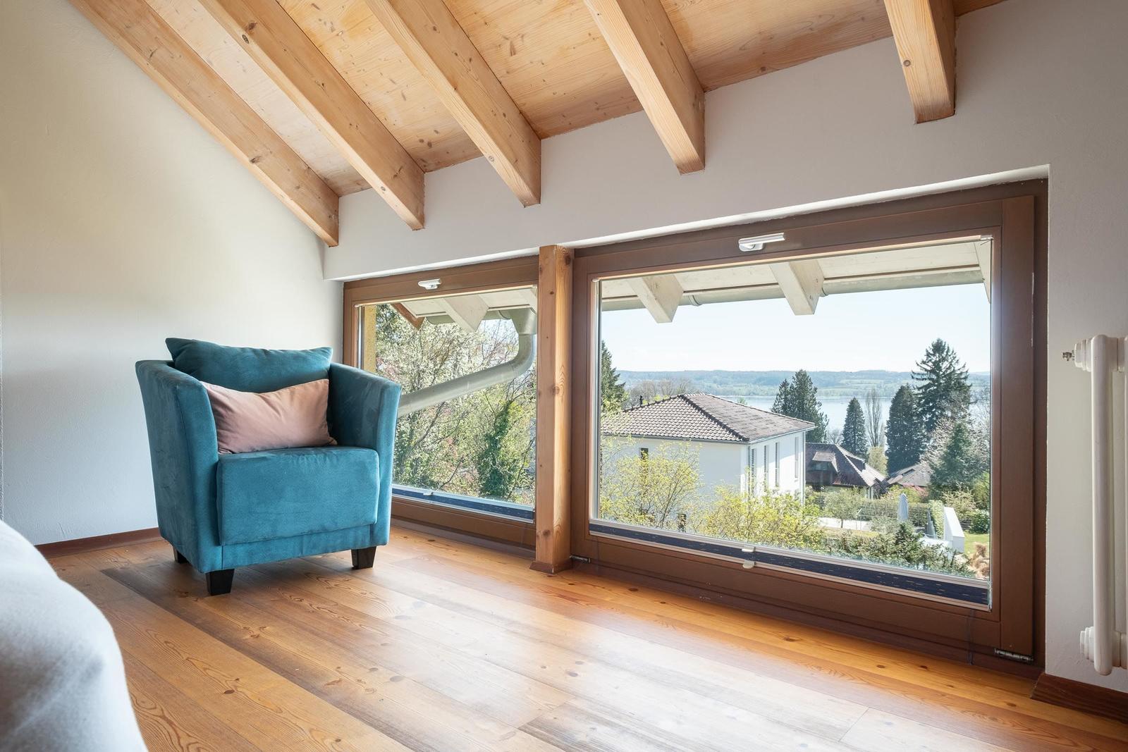 Ein Raum mit Holzboden und Balken. Ein blauer Sessel steht vor großen Fenstern mit Blick auf die Landschaft.