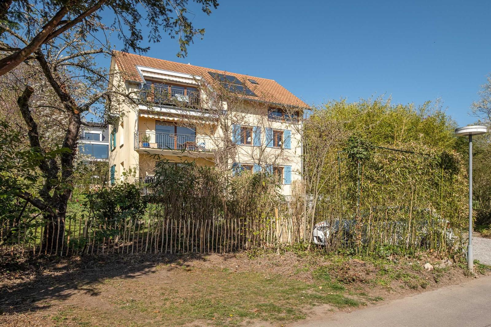 Ein zweistöckiges Haus mit Balkon und Solaranlage, umgeben von Bäumen und einem Zaun.
