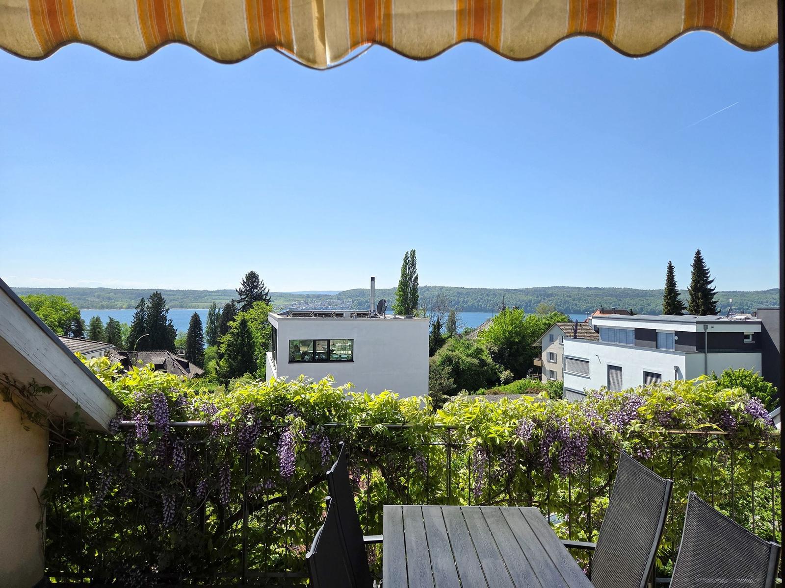 Terrasse mit Tisch und Stühlen, Blick auf See und Häuser unter blauem Himmel.