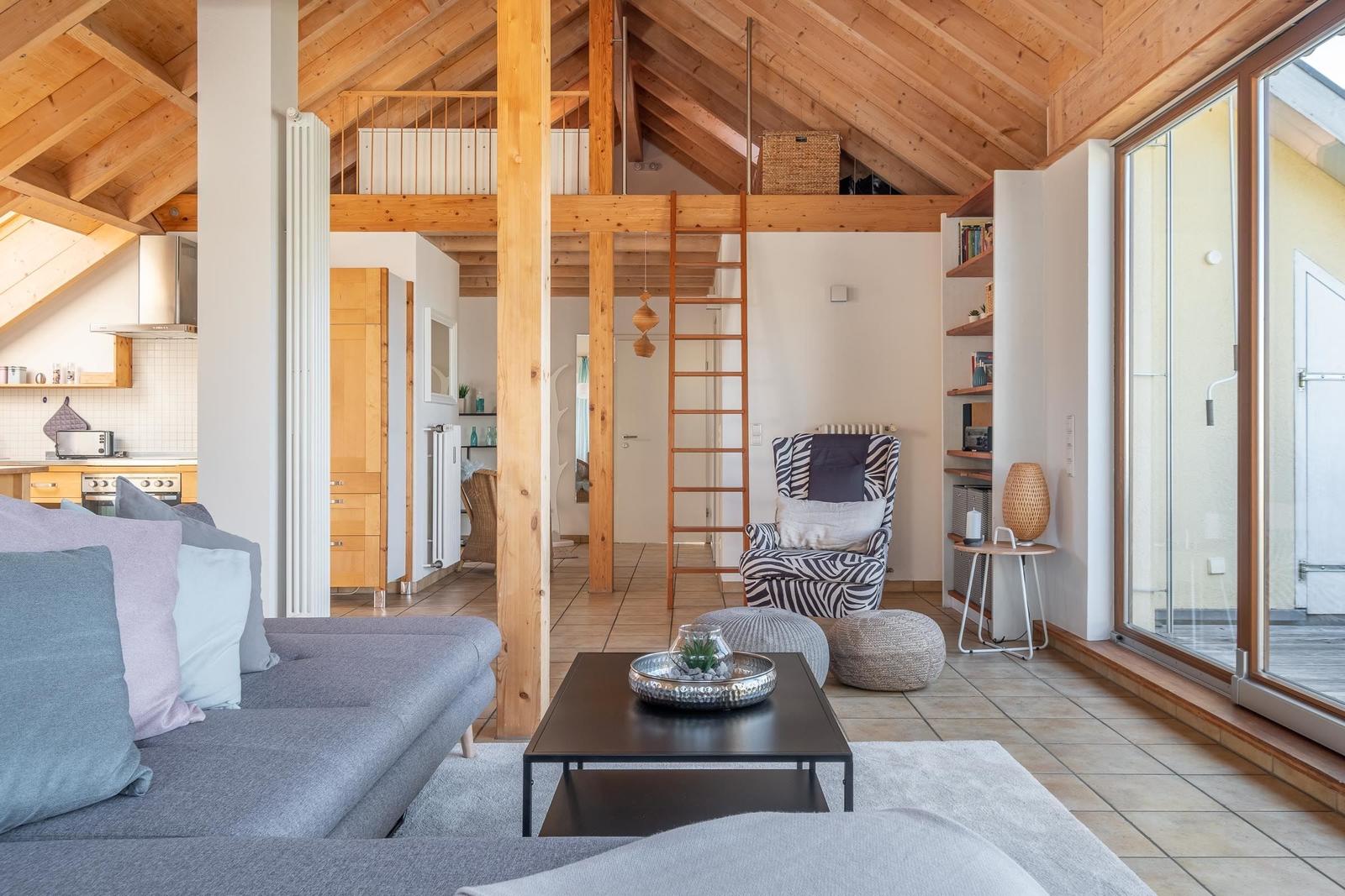 Wohnbereich mit Sofa, Couchtisch und Sessel unter Holzdecke. Treppenhaus und Küchenbereich sichtbar.