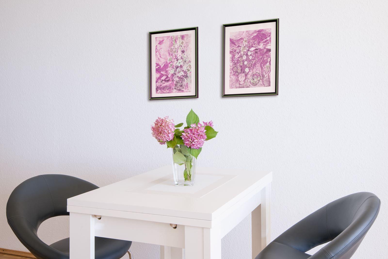 Weißer Tisch mit zwei schwarzen Stühlen und Blumen. Zwei Bilder an der Wand.