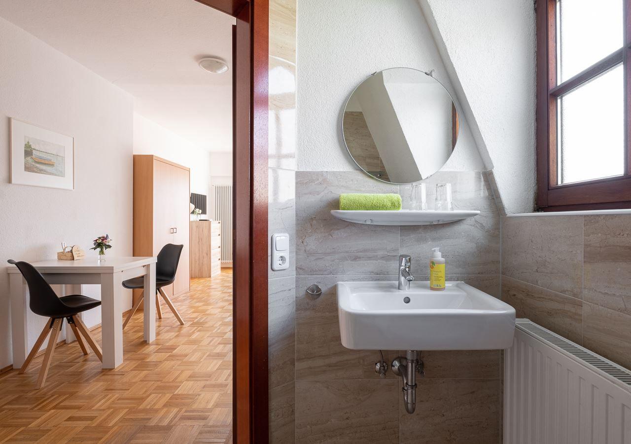 Badezimmer mit Waschbecken, Spiegel und Fenster. Im Hintergrund ist ein Essbereich mit Tisch und Stühlen.