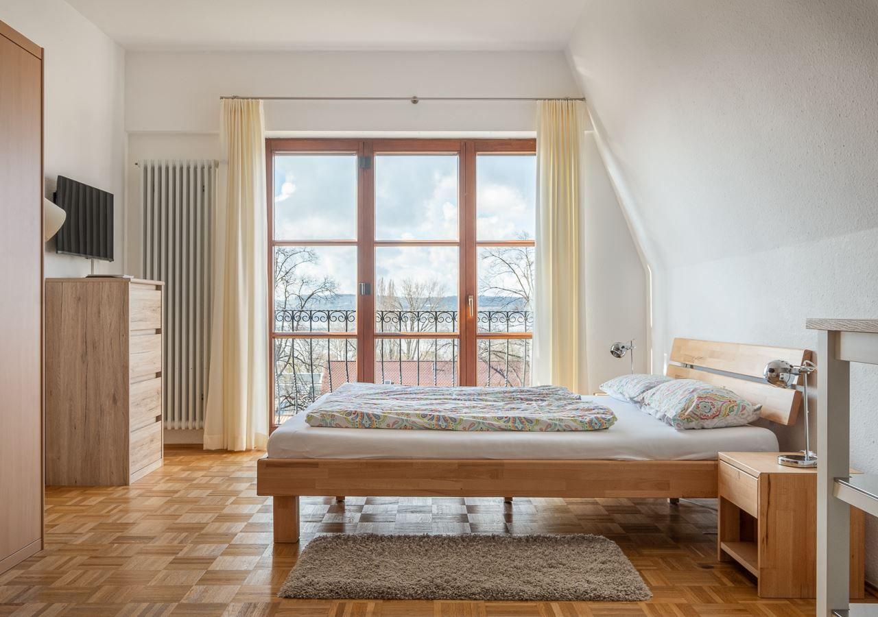Schlafzimmer mit Holzbett, Fenster und Balkon. Parkettboden und Holzmöbel.
