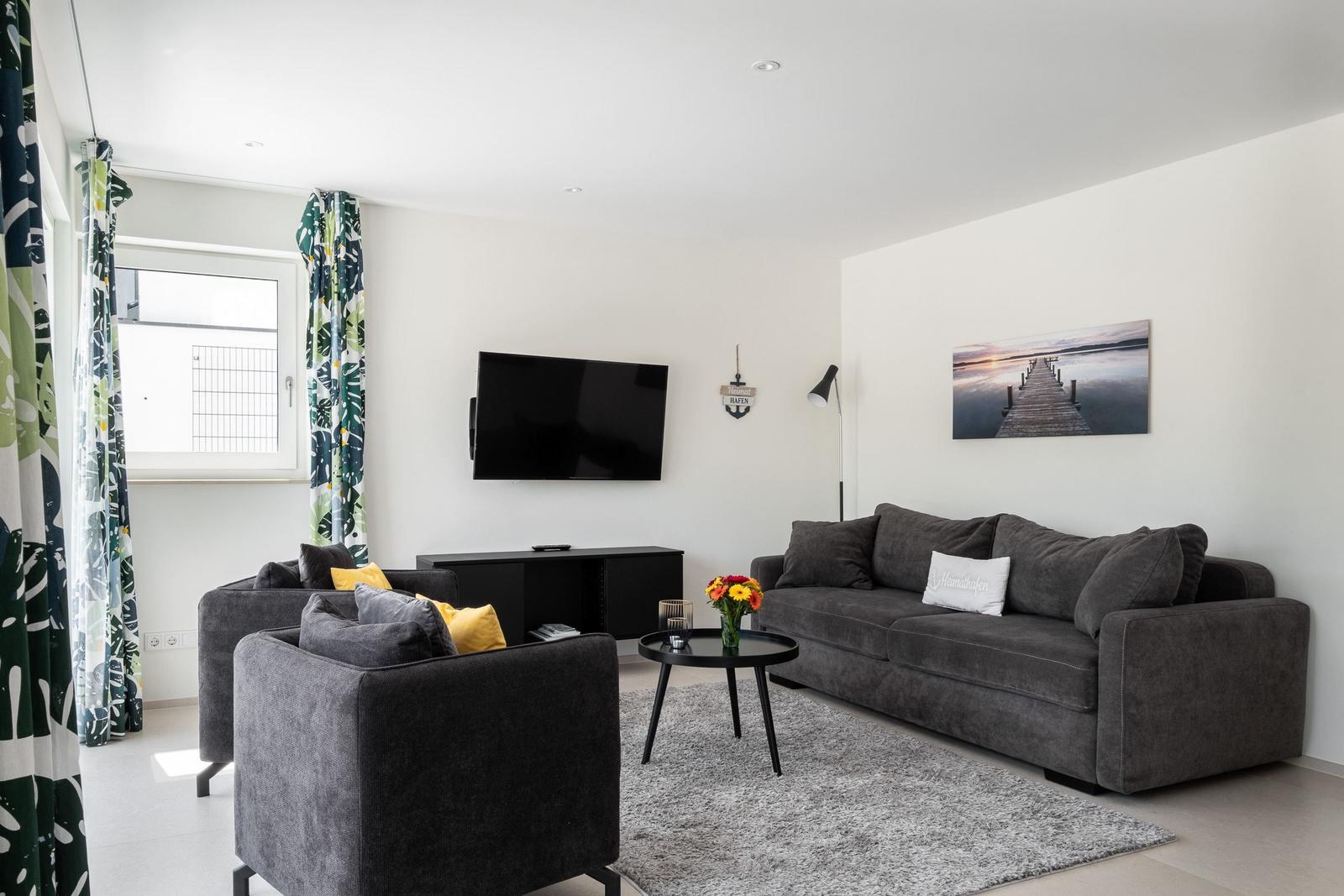 Wohnzimmer mit grauem Sofa, TV und Fenster mit floralen Vorhängen.