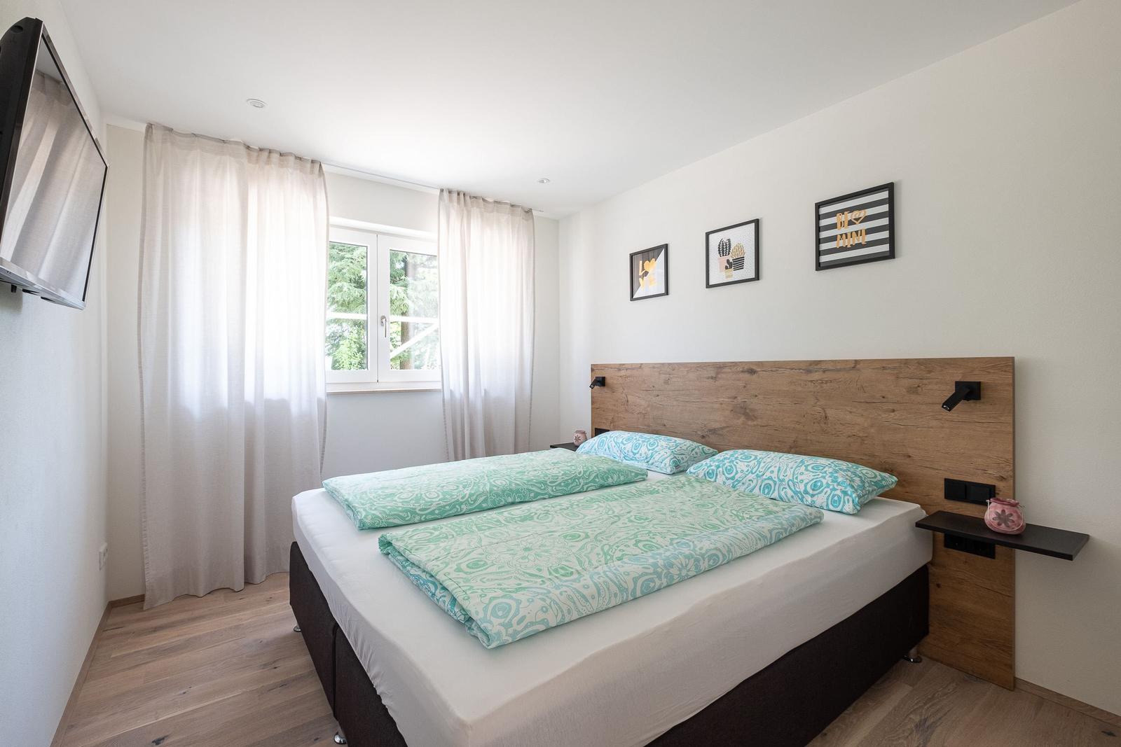 Schlafzimmer mit Doppelbett, Holzkopfteil, Fenster und Wandbildern.