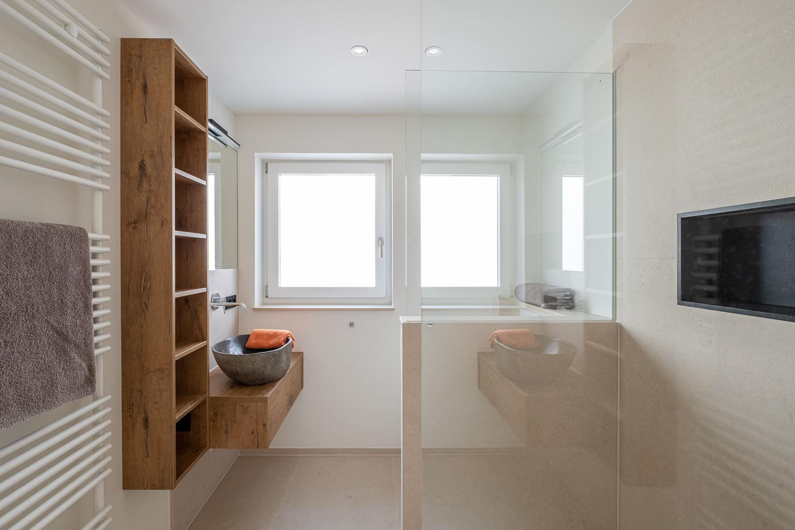 Badezimmer mit Dusche, Waschbecken und Heizung. Holzregal und Fenster sichtbar.