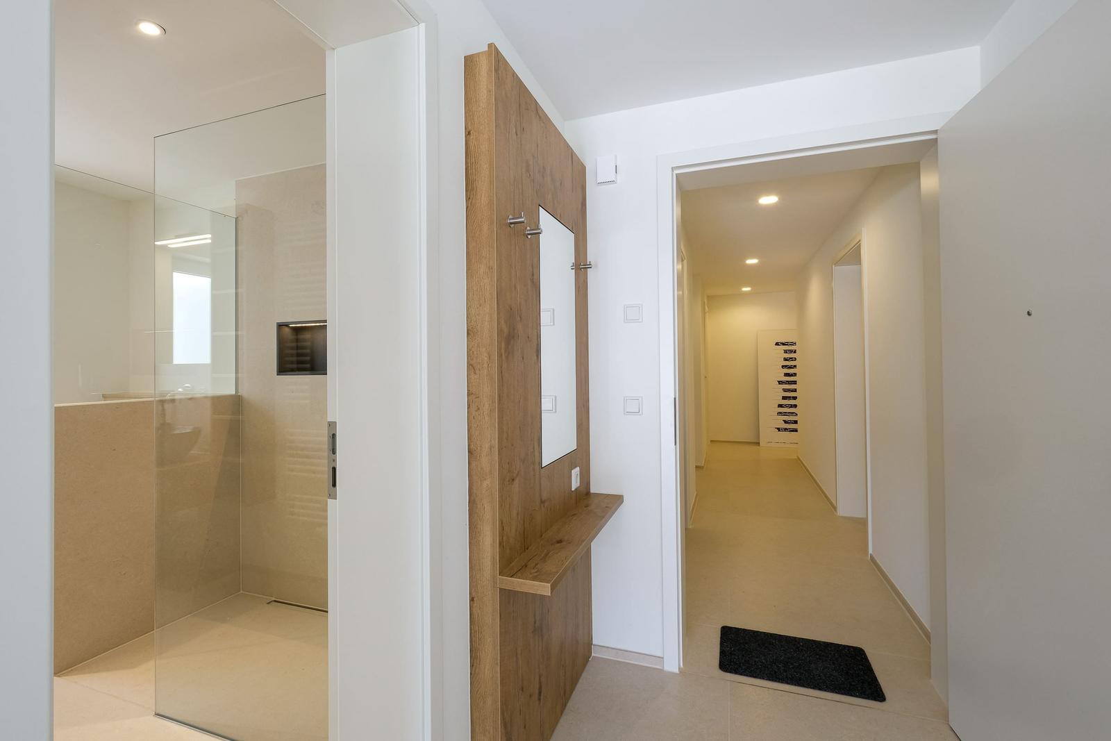 Flur mit Badezimmer und Holzgarderobe. Fliesenboden und schwarzes Teppichchen.