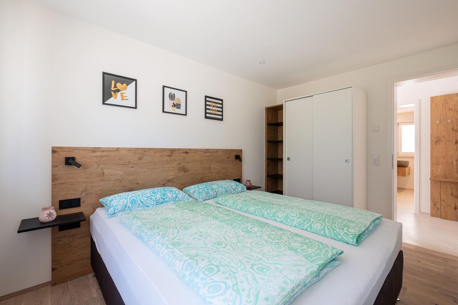 Schlafzimmer mit Doppelbett, Holzkopfteil und Wandbildern. Anschlussbad sichtbar.