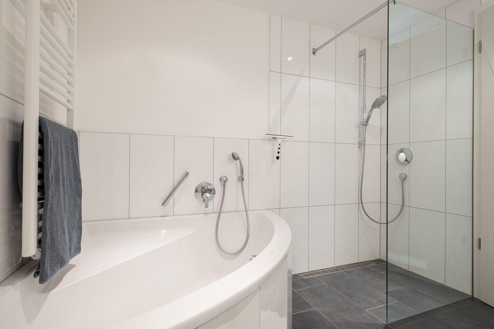 Bad mit Dusche und Whirlpoolbad, weiße Fliesen, graue Bodenfliesen und Handtuchhalter.