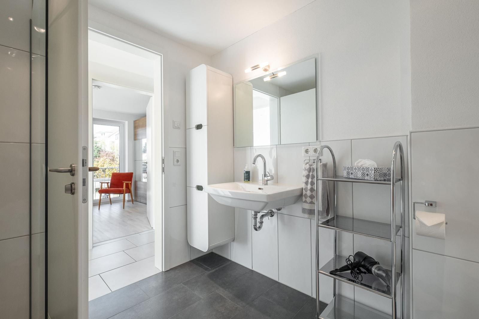 Badezimmer mit Waschbecken, Spiegel und Dusche. Durchgang zu Wohnbereich mit rotem Sessel.