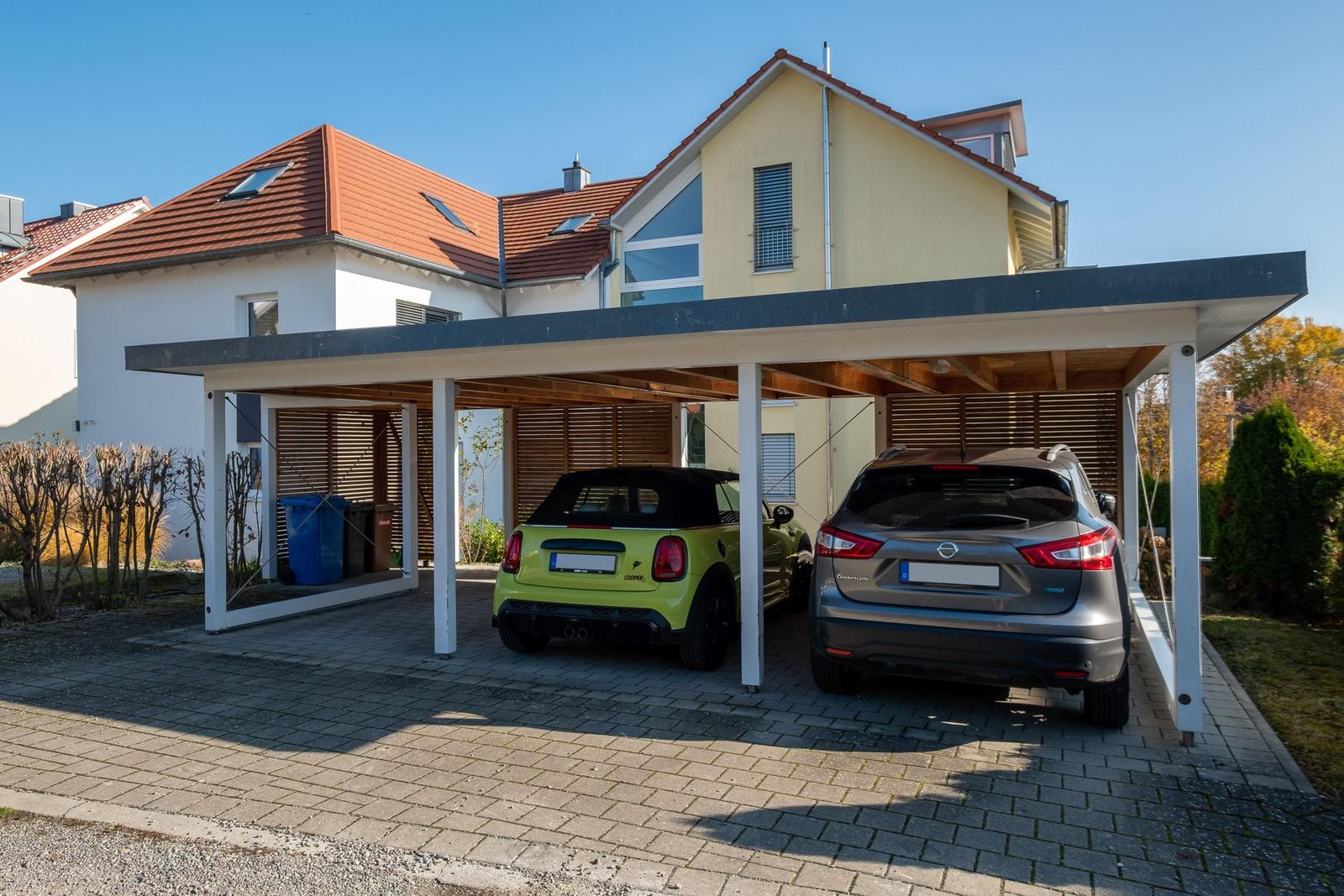 Zwei Fahrzeuge unter einem Carport vor einem Haus mit rotem Dach.