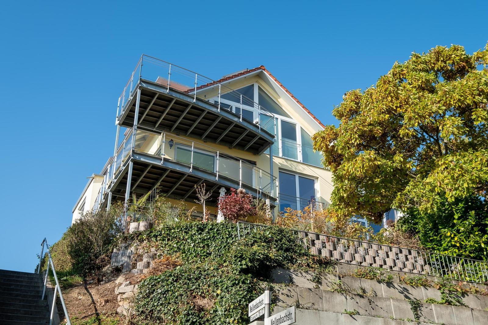 Modernes Haus auf Hang mit Balkonen und Glasfassade. Grün bewachsenes Gelände und Schild Nellenbachstr.