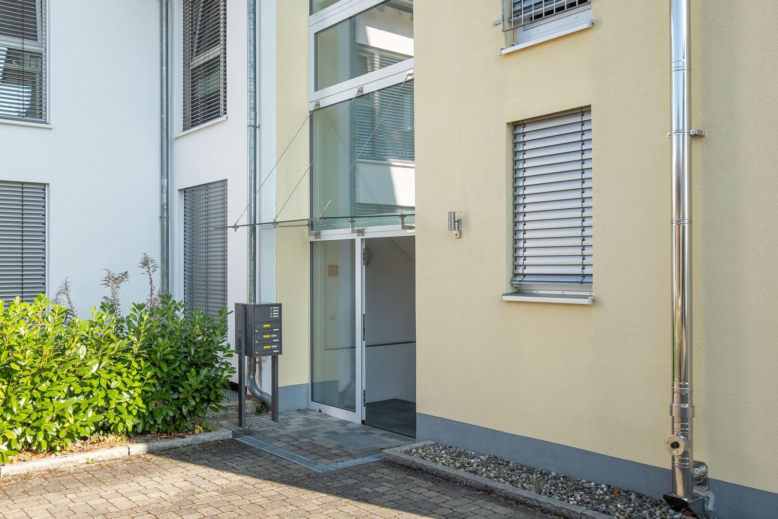 Eingangsbereich mit Glasfront und Briefkasten neben grüner Hecke.