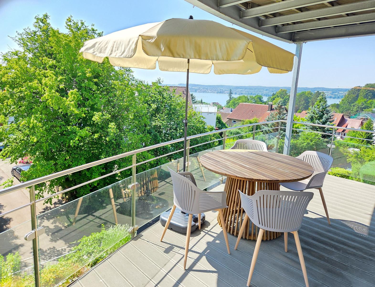 Terrasse mit Tisch, Stühlen und Sonnenschirm. Blick auf Grün und Wasser.