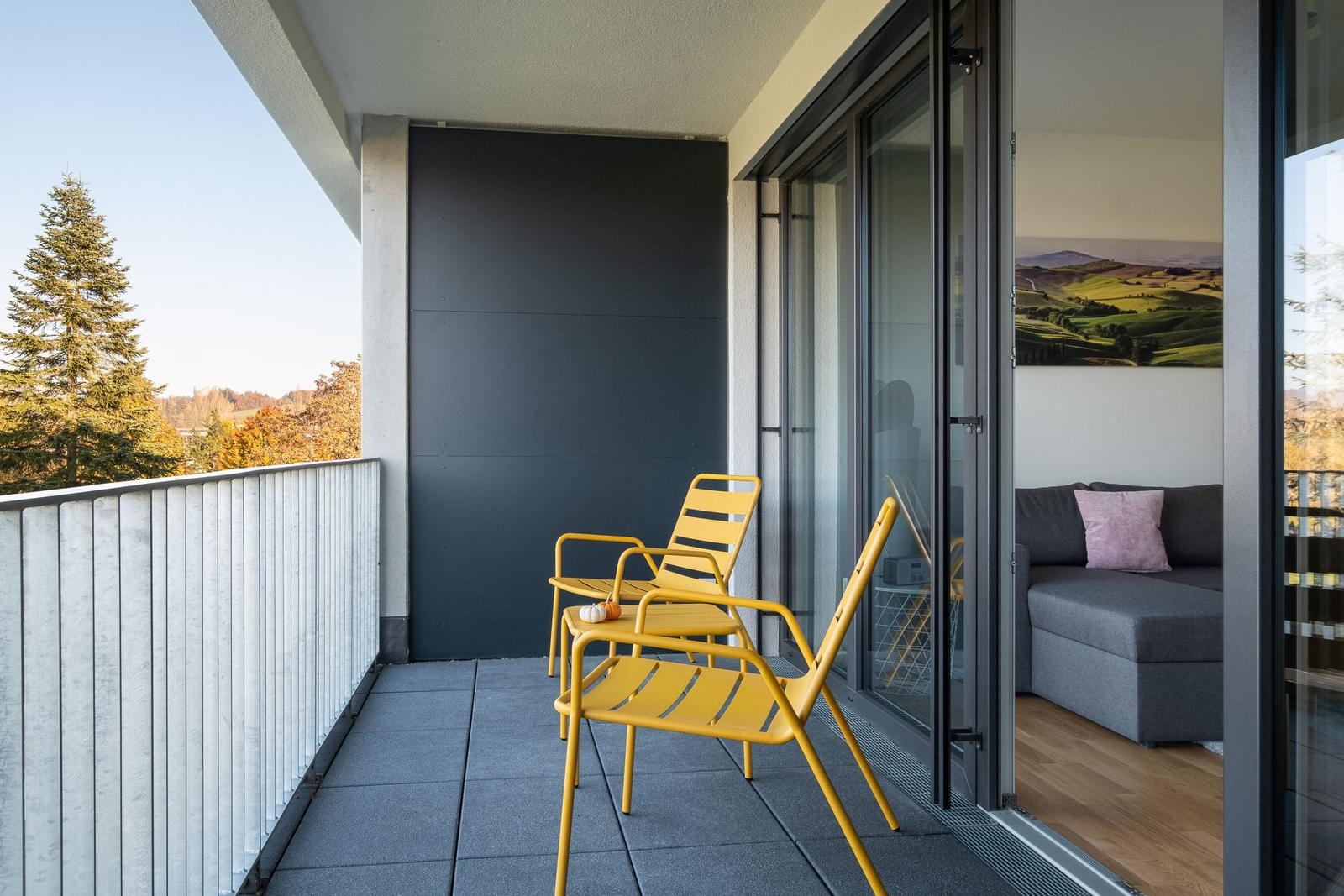 Terrasse mit zwei gelben Stühlen und Blick auf Wald und Hügel. Durchgehende Glastür zum Wohnzimmer.